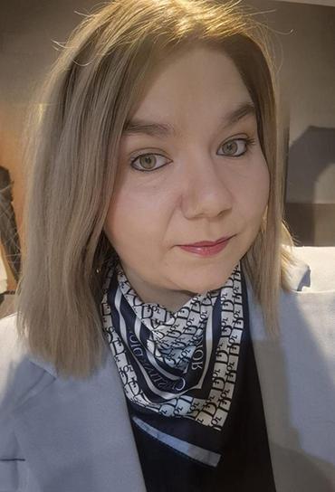 Kobieta o półdługich blond włosach ubrana w czarną bluzkę i szary żakiet oraz wzorzystą apaszkę